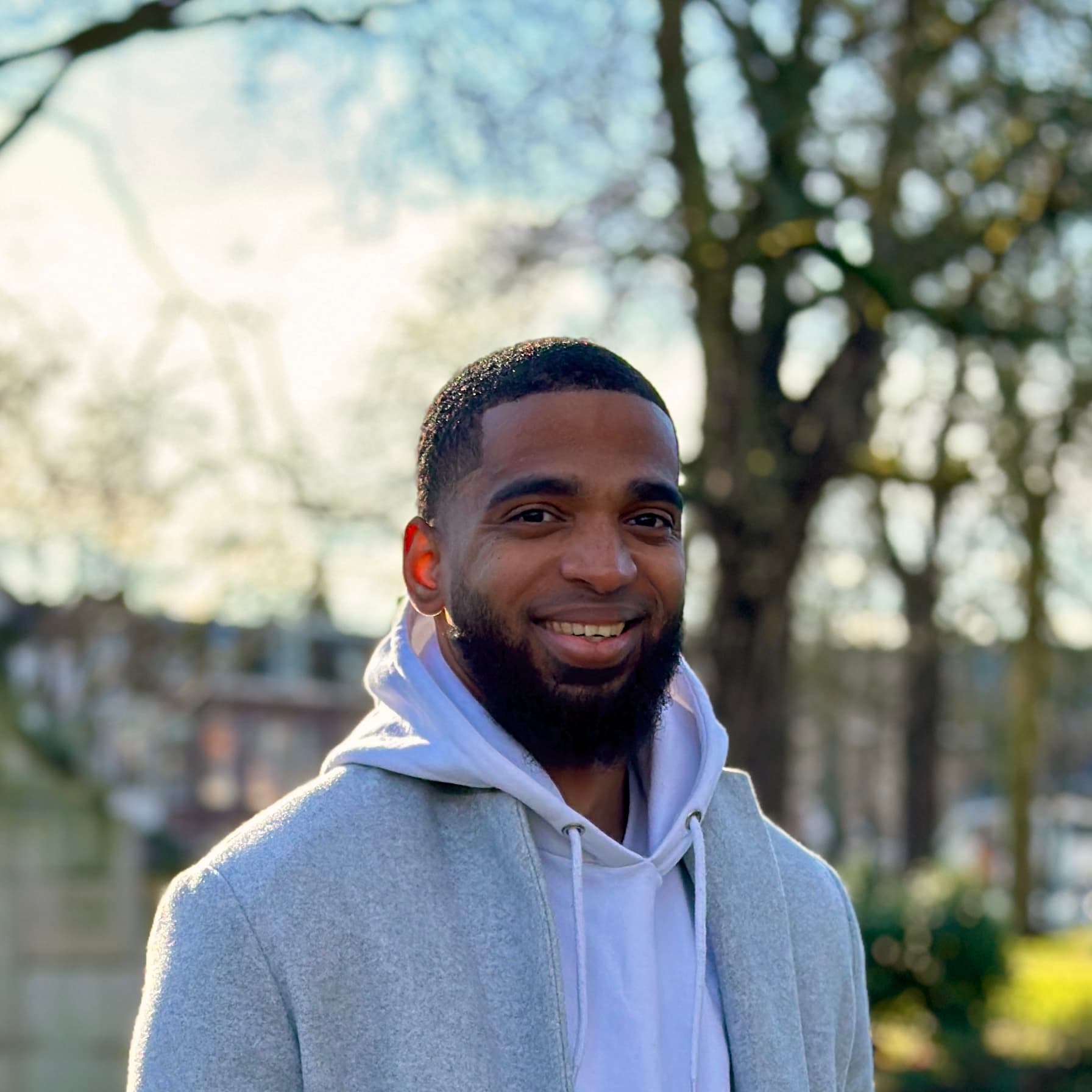 Chris outdoors on a bright day, smiling, in a grey coat over a light hoodie.